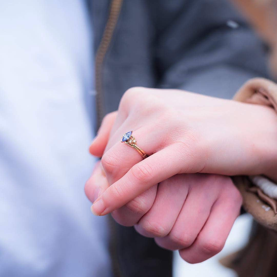 A close-up of a person's hand wearing a gold ring with a blue gemstone, holding hands with another person in a close, intimate moment.