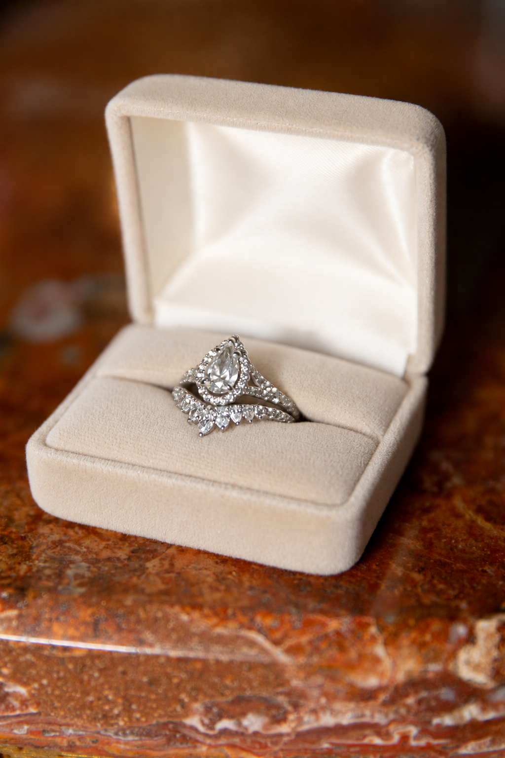 A silver diamond engagement ring with a marquise-cut center stone and pavé band, displayed inside an open beige velvet ring box on a polished brown marble surface.