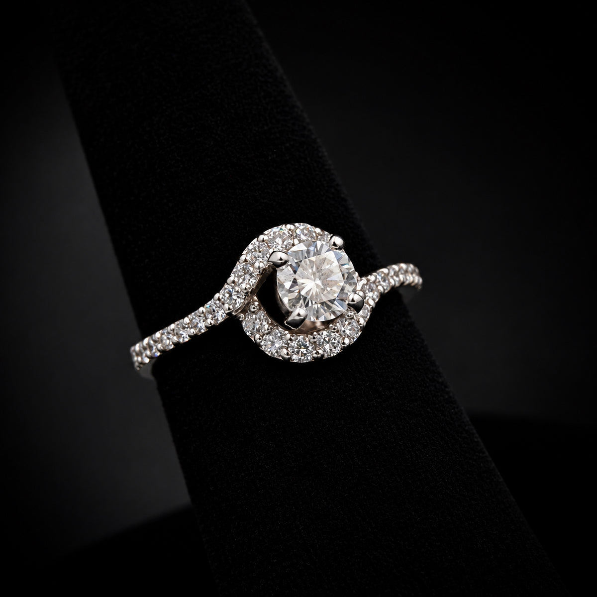 A silver-toned halo ring with a round-cut center stone and pavé-set band, displayed on a black ring stand against a dark background.